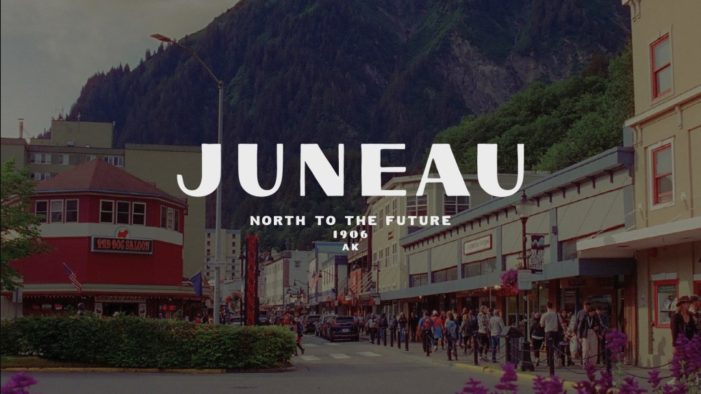 A vibrant street scene in Juneau, Alaska, featuring shops and pedestrians, with mountains in the background. The text overlay reads 'JUNEAU NORTH TO THE FUTURE 1906 AK'.
